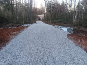 A gravel road under construction in a forested area, with a vehicle and construction materials visible in the distance.