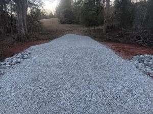 A gravel road extends through a wooded area, bordered by trees and red soil, leading toward an open field in the background.