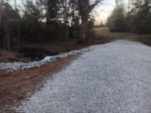 A gravel road curves through a wooded area with trees on both sides and a ditch lined with rocks on the left.