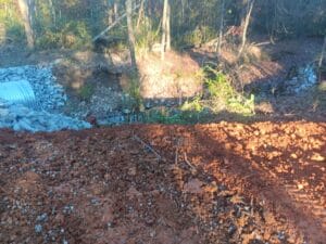 Red dirt and loose soil in the foreground with a small rocky culvert and pipe on the left, leading into a wooded area with trees and sparse vegetation.