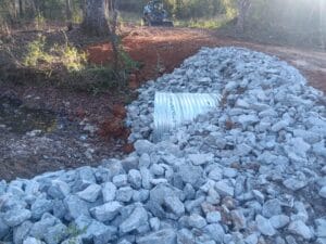 A drainage culvert covered with large rocks is installed under a dirt road in a wooded area, with construction equipment visible in the background.