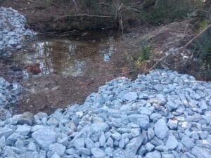 A shallow pool of water surrounded by dirt and vegetation, with two piles of gray rocks in the foreground.