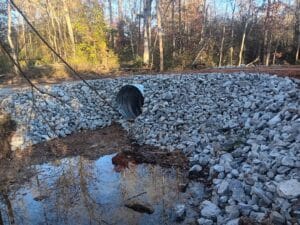 A culvert pipe set in a bank of large gray rocks allows water to flow beneath a dirt roadway in a wooded area.