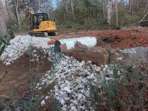 A construction site with a yellow excavator, a large corrugated metal pipe, and piles of rocks and soil in a wooded area.