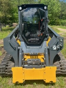 Front view of a John Deere tracked skid steer loader parked on grass, with trees and a dirt path in the background.