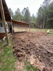 Dirt and uprooted soil cover the ground beside a simple structure with a covered area, surrounded by trees in the background.