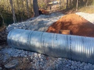 A large metal culvert pipe lies on gravel beside a dirt pile, with construction materials and trees visible along a forested path.
