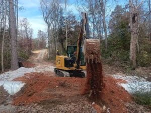 A yellow excavator deposits red dirt over metal culvert pipes at a construction site in a wooded area.