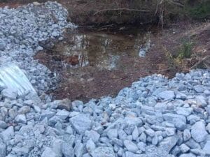 A small, shallow water pit surrounded by piles of gray rocks, with some sparse vegetation and trees in the background.