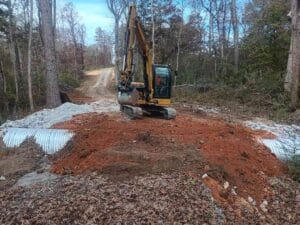 A yellow excavator is parked on a dirt road over large metal culverts, surrounded by trees, with a gravel path extending into the distance.