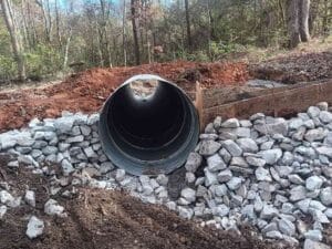 A large metal culvert pipe is installed in the ground with rock and gravel surrounding it, set in a forested area with dirt and trees in the background.