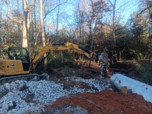 A yellow excavator moves rocks beside a large metal culvert pipe at a construction site near a wooded area.