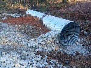 A large corrugated metal culvert pipe is partially buried in gravel and rocks, with dirt piles around it, at a construction site near a wooded area.
