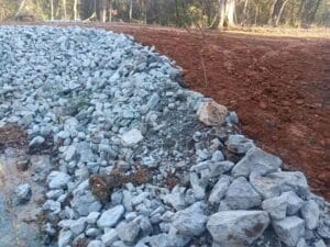 Large gray rocks form a sloped embankment next to bare, reddish soil with trees in the background.