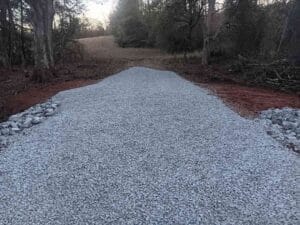 A gravel road under construction runs through a wooded area, with piles of loose stones on either side and trees in the background.