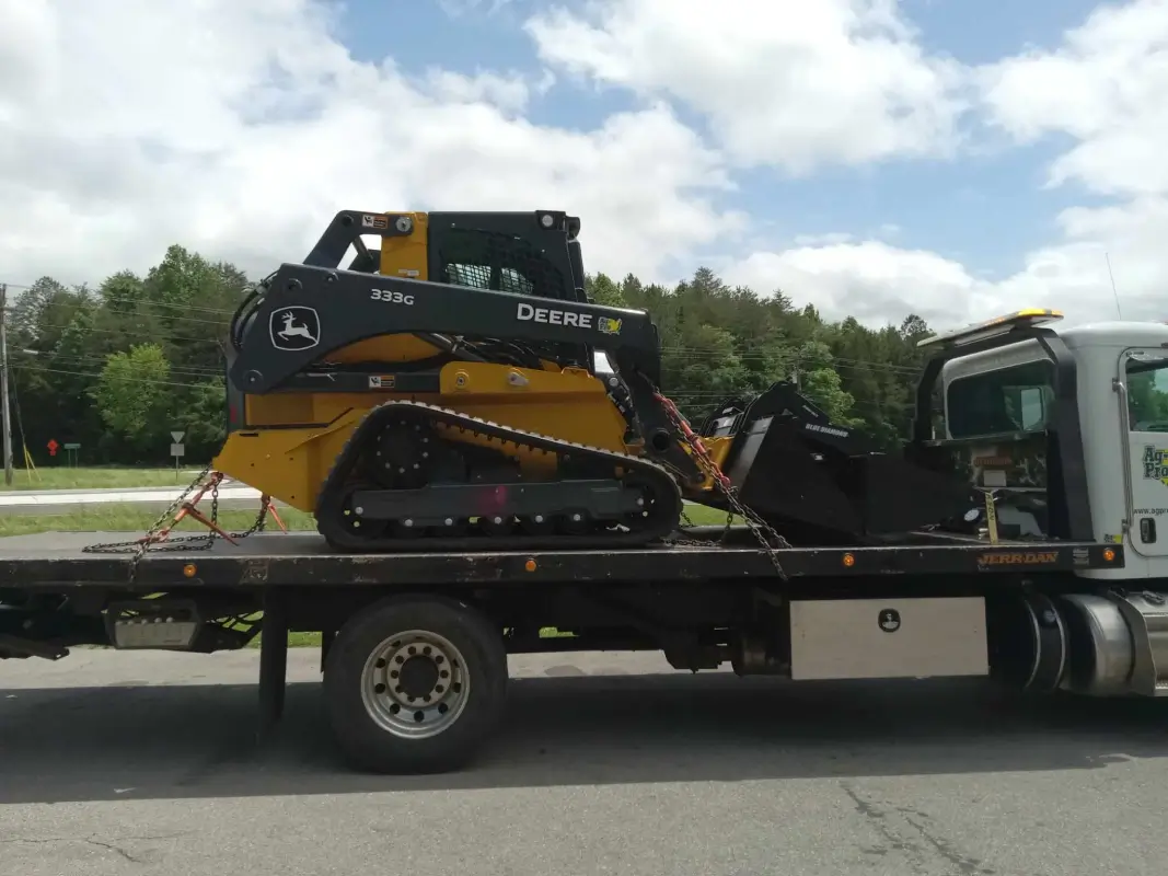 A compact construction vehicle is secured on the flatbed of a truck in an outdoor setting, with trees and a cloudy sky in the background.