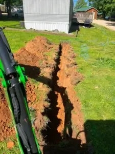 A freshly dug trench runs through a grassy yard near a mobile home, with construction equipment visible in the foreground.