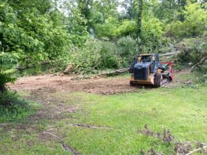 A small bulldozer is clearing fallen trees and brush in a grassy, wooded area.