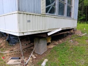 The image shows a corner of a mobile home elevated on concrete blocks, with damaged siding and exposed insulation underneath. Debris and pipes are scattered on the ground nearby.