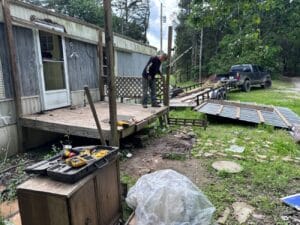 A person repairs a damaged wooden porch next to a mobile home; tools and construction materials are scattered nearby, and a truck with a trailer is parked in the background.