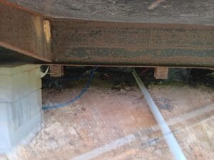 View under a structure showing a metal beam, concrete blocks for support, exposed pipes, wires, and ground covered with dirt and debris.