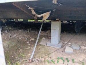 Cinder blocks support a damaged foundation of a building; insulation is exposed from a tear in the floor above, and debris is scattered on the ground.