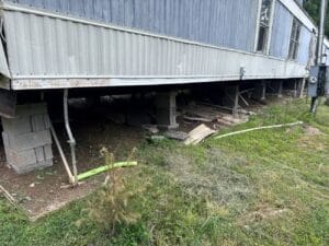 The underside of a mobile home elevated on concrete blocks, with exposed dirt, scattered wood, pipes, and overgrown grass in the surrounding area.