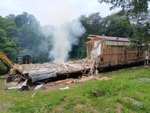 A partially demolished building with debris and smoke rising from the remains, surrounded by trees and grass, with an excavator visible on the left.