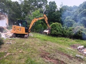 A person operates a yellow excavator to clear debris near a house, with trees and bushes in the background.