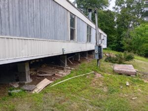A mobile home elevated on concrete blocks with exposed underside and scattered debris, set in a grassy yard with trees in the background.
