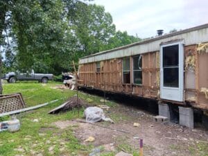 A mobile home with exterior walls partially removed sits elevated on blocks, exposing insulation and framing; debris and tools are scattered nearby.