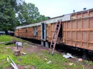 A mobile home with its exterior siding removed, exposing insulation and framing, stands on concrete blocks amidst construction materials and debris.