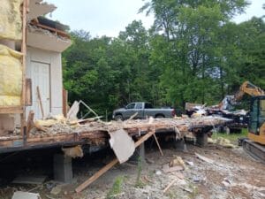 Partially demolished building with exposed insulation and debris; construction equipment and a pickup truck are visible nearby among trees.