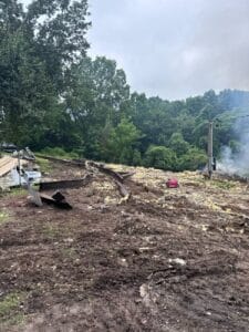 A burned and demolished building site with scattered debris, smoke rising, exposed beams, and surrounding green trees under an overcast sky.