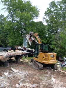 A yellow CAT excavator is demolishing a building, lifting debris on a construction site surrounded by trees.