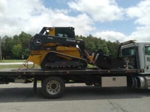 A John Deere 333G compact track loader is secured on the bed of a flatbed tow truck, parked on a paved road with trees and cloudy sky in the background.