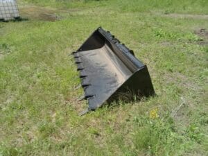 A black metal loader bucket attachment is lying on grassy ground outdoors on a sunny day.